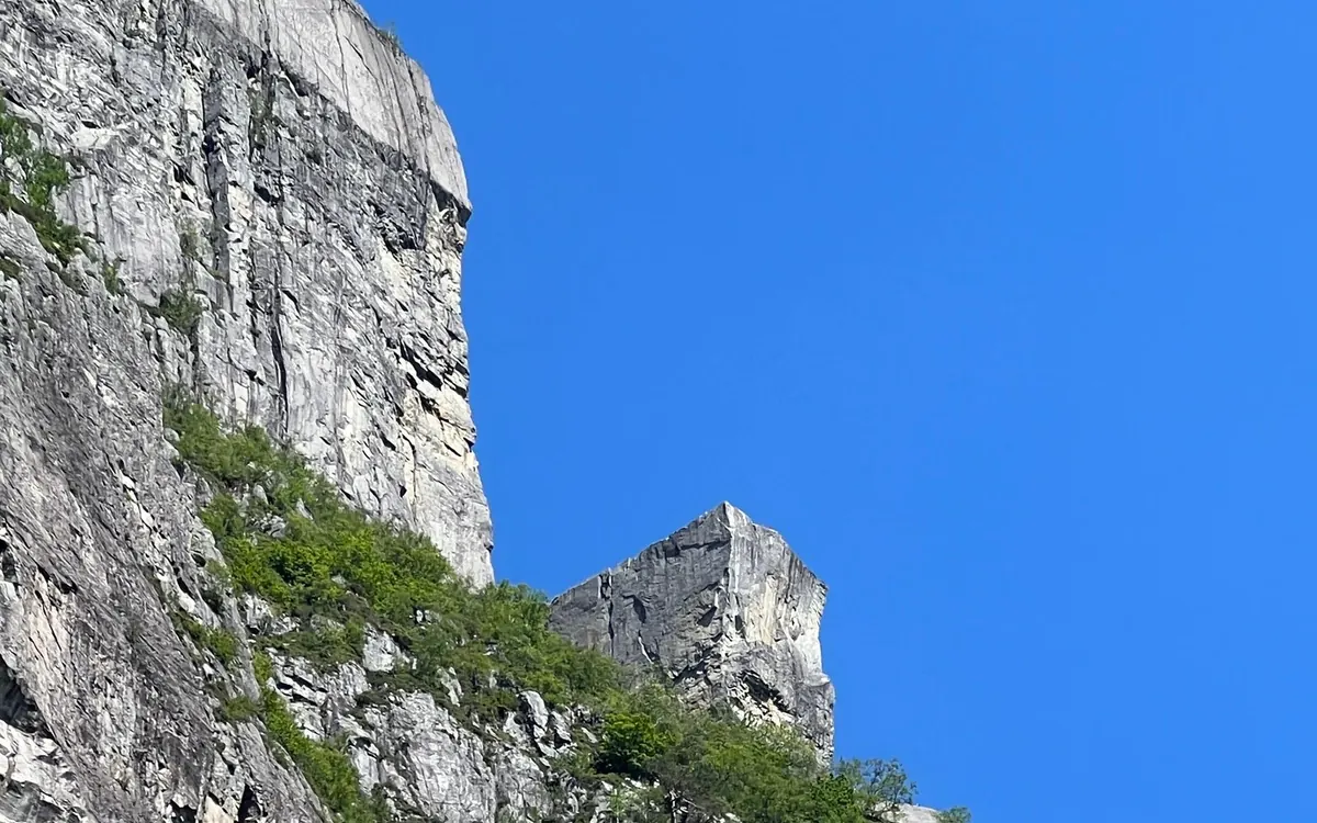 A square granite ledge juts out from a sheer cliff into a deep blue sky, with green scrub below