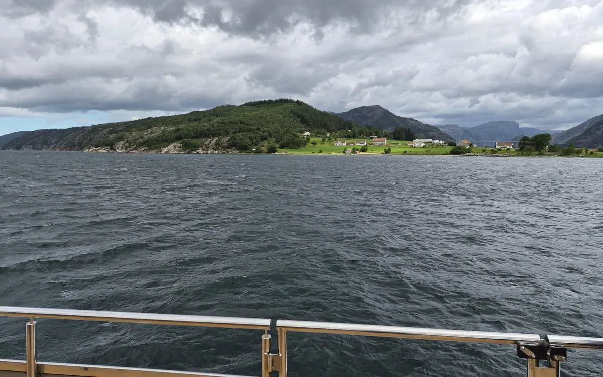 A steel deck railing in front of choppy grey water, white farmhouses on a green shore