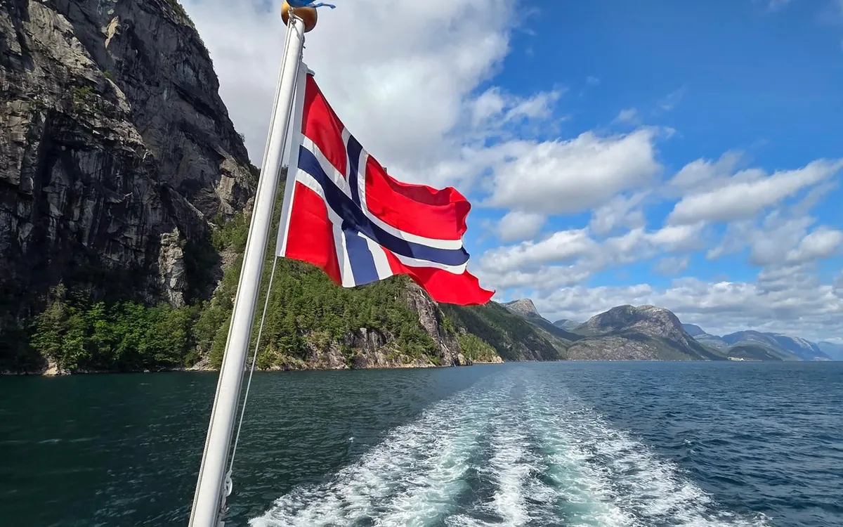 A flag flying above the white wake behind a ship on the fjord