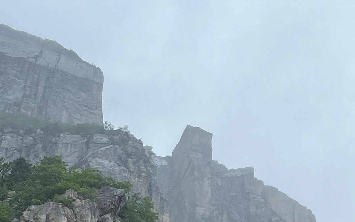 A sheer pale wall and a square-cut block in grey haze, trees on the rocks below