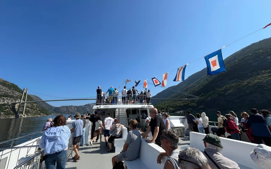 Passengers on the open deck of a large white boat, signal flags strung overhead, steep hills either side