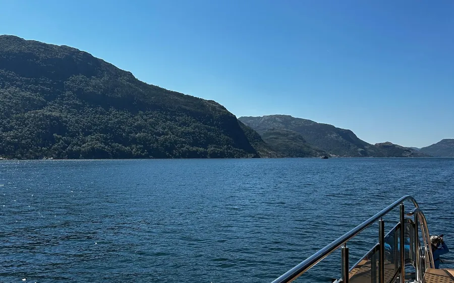 The rail of a boat above deep blue water, forested hills on both shores