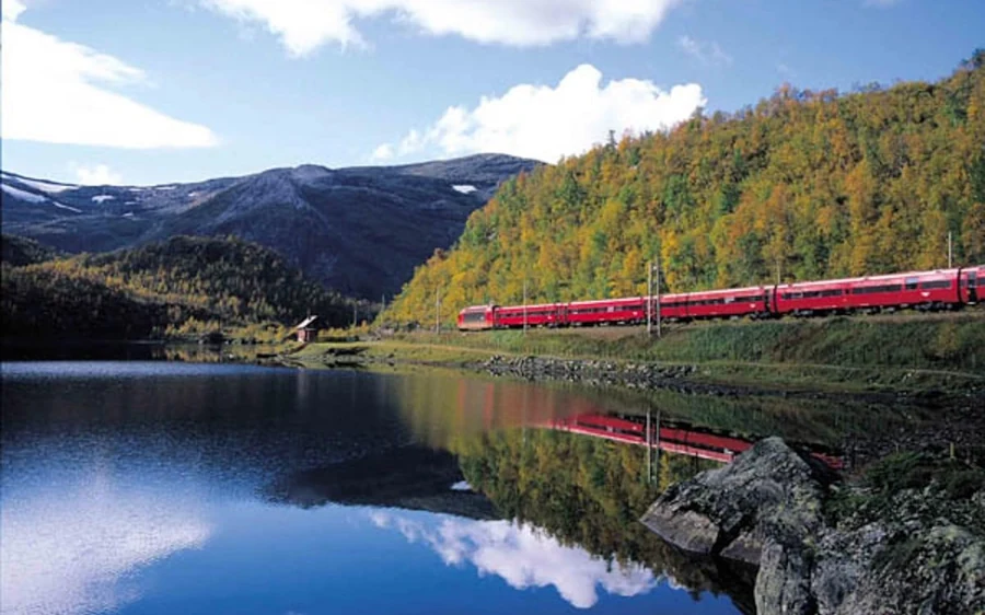 A red train passing autumn trees beside a still lake that mirrors the hills