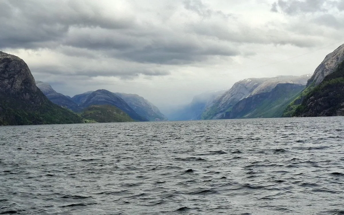 A wide grey fjord under heavy cloud, with steep green and grey mountains on both sides